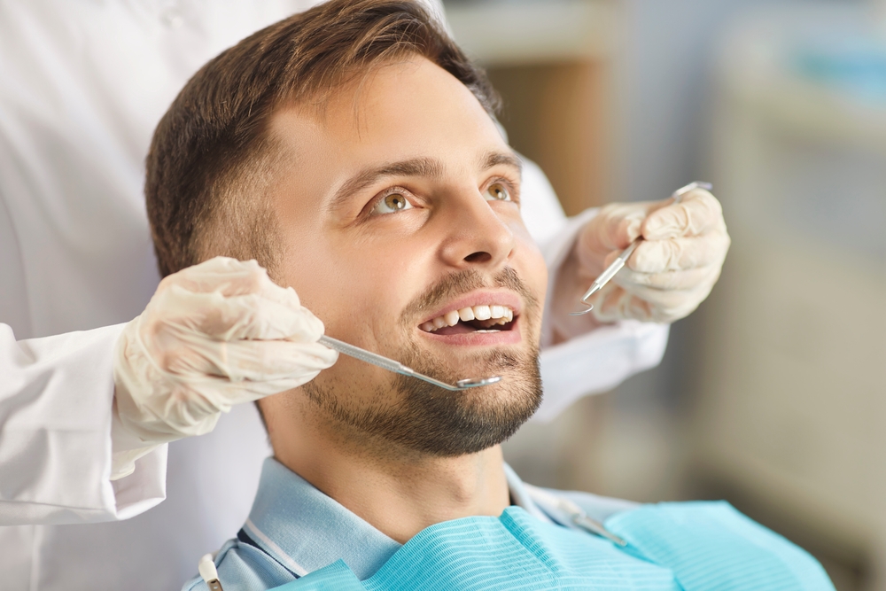 Patient at Dental Checkup - Volterra Dental Comprehensive and Aesthetic Dentistry Male patient smiling during a dental checkup with dentist using dental instruments. - Volterra Dental Comprehensive and Aesthetic Dentistry